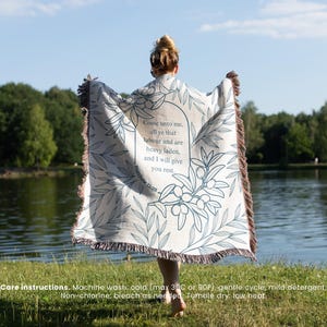 May include: A cream-coloured throw blanket with a botanical design and text. The blanket features a quote in a stylized font, surrounded by olive branches and leaves. The edges have a brown fringe.