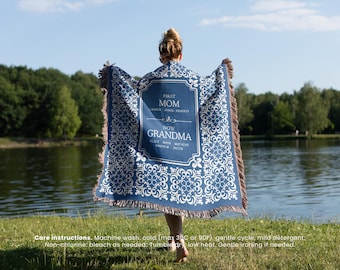 May include: A blue throw blanket with a white floral design and brown fringe. The blanket has the text "FIRST MOM NOW GRANDMA" with names below. The blanket is held up by a person near a lake.