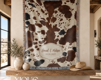 May include: A woven throw blanket featuring a brown, black, and white cowhide print pattern with fringed edges. A central cream-colored decorative label displays the text Daniel & Melissa and the date 10.20.2027 in a brown script and serif font.