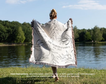 May include: A white throw blanket with a black olive branch design and the text "THE MARTIN FAMILY Est. 2023". The blanket has a fringe border and is draped over a person's shoulders.