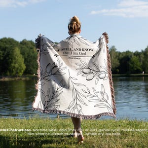 May include: A cream-colored throw blanket with a black olive branch design and the text "BE STILL, AND KNOW that I am God, PSALM 46:10." The blanket has a fringed edge and is held up by a person standing near a lake.