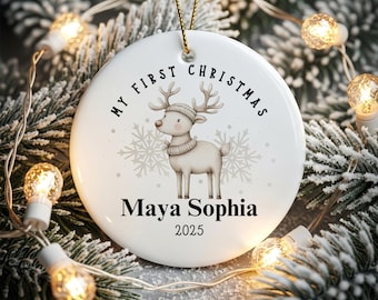 May include: A white ceramic Christmas ornament with the text "MY FIRST CHRISTMAS" and the name "Maya Sophia" below a cartoon reindeer wearing a hat and scarf. The year "2025" is printed below the name. The ornament hangs from a gold string.