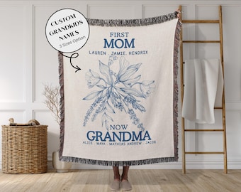May include: Cream-colored throw blanket with blue floral design and fringe. Text reads "First Mom" and "Now Grandma" with names below. The blanket is displayed in front of a wooden ladder and a wicker basket.