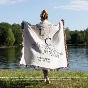 Può includere: Una coperta personalizzata color crema con un motivo floreale decorativo e le iniziali "L & C". La coperta ha un bordo con frange e il testo "THE SLACKS July 24th, 2025". La coperta è tenuta davanti a un lago.
