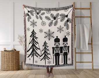 May include: A cream-colored woven throw blanket with a black Christmas-themed design. The design includes snowflakes, Christmas trees, ornaments, and nutcracker soldiers. The blanket has a fringed edge.