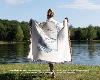 May include: A white throw blanket with a blue floral design and text that reads "First Mom Now Grandma." The blanket has a fringed edge and is being held up in front of a lake.