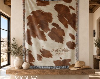 May include: A woven throw blanket featuring a brown and white cowhide pattern with fringed edges. The blanket is personalized with the text 'Daniel & Melissa' and the date '10.20.2027' printed in a brown script font on the lower right side.