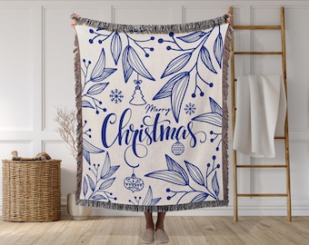 May include: A cream-colored throw blanket with a blue floral design and the words "Merry Christmas" in a script font. The blanket has a fringed edge and features Christmas ornaments and snowflakes.
