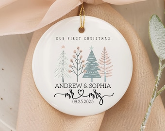 May include: A white ceramic Christmas ornament with a gold string. The ornament features the text "OUR FIRST CHRISTMAS" above four illustrated trees. Below the trees are the names "ANDREW & SOPHIA", the words "mr & mrs" with a heart, and the date "09.25.2025".
