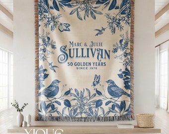 May include: A woven throw blanket featuring a cream background with blue botanical illustrations of birds, butterflies, and berries. The text reads Marc & Julie Sullivan 50 Golden Years Since 1976. The blanket has a fringed edge with multicolored threads.