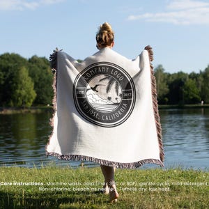 May include: A cream-colored blanket with a black and white circular logo featuring a palm tree and the text "COMPANY LOGO SAN JOSE, CALIFORNIA ESTD 2017". The blanket has fringed edges and care instructions at the bottom.