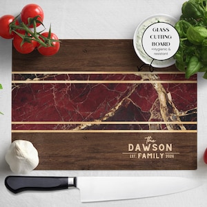 May include: A rectangular wooden cutting board with a dark brown border and a central section featuring a red marble design with gold accents. The text "The Dawson Family Est. 2026" is engraved on the board. A white bowl with a green dip, tomatoes, basil, garlic, and a knife are next to the board.