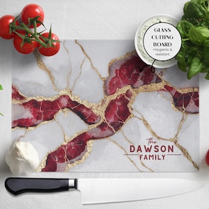 May include: A rectangular glass cutting board with a white marble design, accented with red and gold veining. The text "The Dawson Family" is printed in burgundy. The board is surrounded by fresh basil, tomatoes, and a knife.
