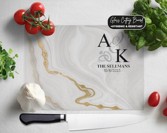 Może przedstawiać: Prostokątna szklana deska do krojenia z białym i złotym marmurowym wzorem. Deska zawiera tekst "Glass Cutting Board +HYGIENIC & RESISTANT" oraz spersonalizowany tekst. Obecny jest również nóż szefa kuchni, pomidory, bazylia i czosnek.