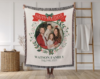 May include: Cream-colored woven throw blanket with a family photo surrounded by a green wreath. The blanket has a red banner that reads "HAPPY HOLIDAYS" and the text "WATSON FAMILY July 24th, 2025". The blanket has a fringe.