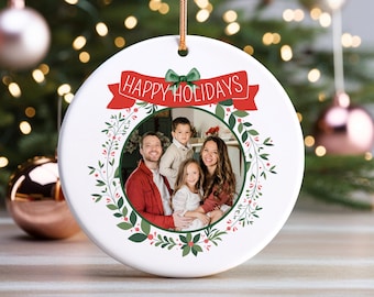 May include: White ceramic Christmas ornament with a family photo inside a green wreath. The ornament has a red banner with the words "HAPPY HOLIDAYS" and a green bow. A gold string is attached for hanging.