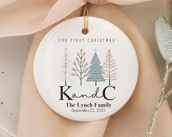 May include: White ceramic Christmas ornament with a gold string. The ornament features four stylized Christmas trees in pastel colors and the text "OUR FIRST CHRISTMAS". Below the trees is the name "KandC" and the text "The Lynch Family September 25, 2025".