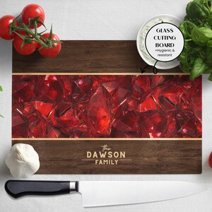 Può includere: Un tagliere rettangolare in vetro con un bordo in legno scuro e un design centrale di pietre preziose rosse. Il testo "The Dawson Family" è stampato in basso. La tavola è etichettata "Glass Cutting Board + Hygienic & resistant".