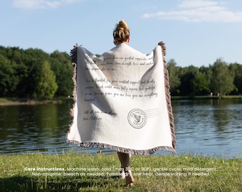 May include: A cream-colored throw blanket with dark brown fringe is held up by a person. The blanket has printed text and a circular stamp design. The text includes phrases like "greatest supporter" and "you are love, you are everything."