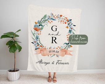 May include: Cream-colored blanket with a floral wreath design. The wreath surrounds the initials "G and R" and the date "07.10.22". The words "Always & Forever" are written below. A small green tag says "Pick from 3 sizes/5 colors".