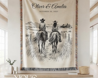 May include: A cream-colored woven throw blanket featuring a black line art illustration of two people on horseback in a rural landscape. The text reads Oliver & Amelia and OCTOBER 20, 2027. The blanket has a multicolored fringed edge on all sides.