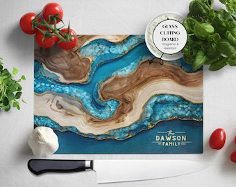 May include: A rectangular glass cutting board with a blue, brown, and gold abstract design. The board has the text "The Dawson Family" and "Est. 2021". A white bowl, tomatoes, basil, garlic, and a knife are next to the board.