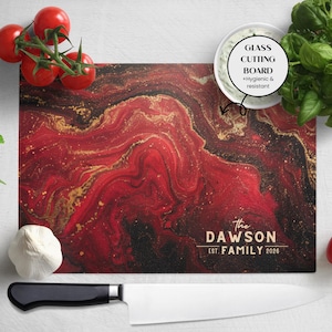 Può includere: Tagliere rettangolare in vetro con un design astratto rosso, nero e oro. Il testo "The Dawson Est. FAMILY 2026" è stampato sulla tavola. Un coltello, pomodori, basilico e aglio sono disposti intorno.