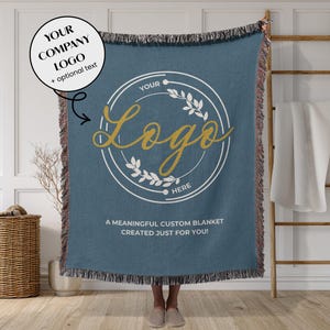 May include: A blue woven blanket with a custom logo design. The blanket features a circular logo with text and a floral design. The text on the blanket reads "YOUR LOGO HERE" and "A MEANINGFUL CUSTOM BLANKET CREATED JUST FOR YOU!"