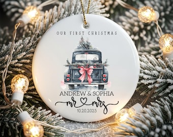 May include: White ceramic Christmas ornament with a black truck carrying a Christmas tree and a pink bow. The text "OUR FIRST CHRISTMAS" is at the top, with names and a date below. A gold string is attached for hanging.