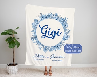 May include: A cream-colored blanket with a blue floral wreath design. The word "Gigi" is in the center, surrounded by a floral wreath. Below the wreath are the words "Mother • Grandma" and dates. The blanket has a tag that says "Pick from 3 SIZES/5 COLORS."