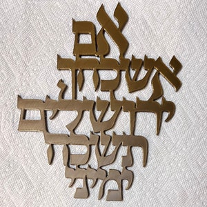 May include: A gold metal wall decor featuring Hebrew lettering. The letters are arranged in a decorative pattern and are raised from the surface.