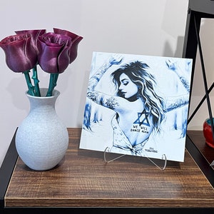 May include: A white canvas print with a blue and white illustration of a woman with tattoos. The text "We will dance again" is printed on the canvas. The print is on a clear acrylic stand and is on a wooden shelf. A bouquet of purple artificial roses is in a white vase on the shelf.