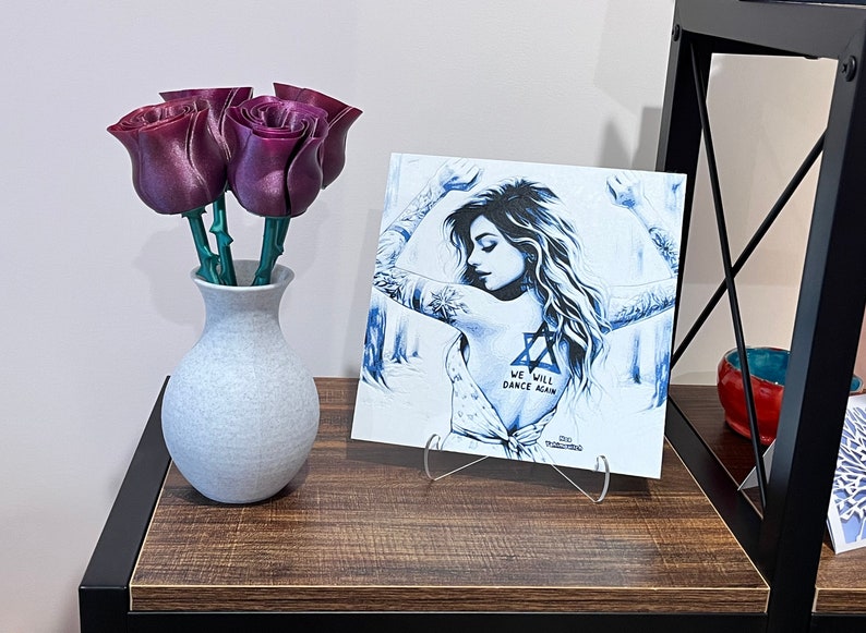May include: A white canvas print with a blue and white illustration of a woman with tattoos. The text "We will dance again" is printed on the canvas. The print is on a clear acrylic stand and is on a wooden shelf. A bouquet of purple artificial roses is in a white vase on the shelf.