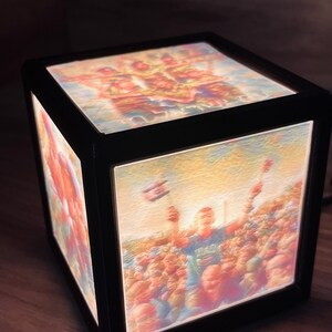 May include: A black cube-shaped night light with a clear front panel displaying a colorful image of a crowd of people. The image is illuminated from behind, creating a soft glow.