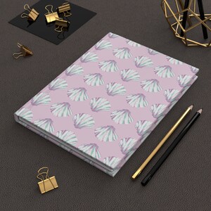 May include: A pink notebook with a white and green seashell pattern. The notebook is closed and lying on a brown leather surface with two pencils and a gold metal stand.
