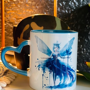 May include: A white ceramic mug with a blue rim and handle. The mug features a watercolor illustration of a blue fairy with wings and a flower. The handle is shaped like a heart.