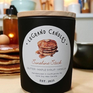 Könnte beinhalten: Schwarze Kerze mit Holzdeckel. Das Etikett zeigt eine Pfannkuchen-Illustration und den Text "Sunshine Stack". Das Etikett listet auch "Butter, Ahornsirup, Vanille" und "EST. 2023" auf.