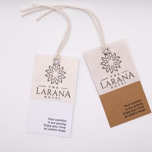 Könnte beinhalten: Zwei rechteckige Anhänger mit dem Text "THE LARANA HOTEL" und einem floralen Design. Die Anhänger sind weiß und beige mit einem angebrachten Faden. Der Text "Your comfort is our priority. Enjoy your time at Larana Hotel" ist unten aufgedruckt.