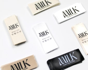 May include: A collection of fabric labels in various colors, including beige, white, and black. Each label features the word "MILK" in a stylized font, along with "CREATIVE STUDIO" and fabric care symbols. The labels are made of a soft, woven material.