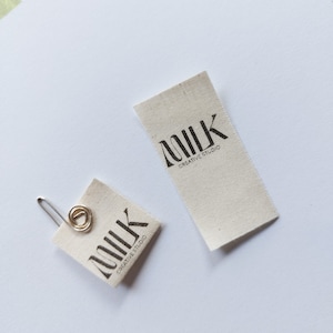 Puede incluir: Dos etiquetas de tela blanquecina con la palabra "MILK" en una fuente negra estilizada. Las palabras "CREATIVE STUDIO" están debajo de la palabra "MILK". Una etiqueta está sujeta a un clip plateado. Las etiquetas están sobre un fondo blanco.