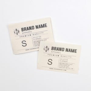 Puede incluir: Dos etiquetas de ropa blanquecinas con texto negro. Las etiquetas dicen "BRAND NAME APPAREL PREMIUM QUALITY" e incluyen la talla "S". Las etiquetas también enumeran la composición de la tela y las instrucciones de cuidado.