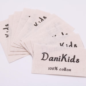 Può includere: Un gruppo di etichette in tessuto beige con la scritta "DaniKids" e "100% cotone" stampata in nero. La parola "kids" è stampata anche su diverse altre etichette. Le etichette sono disposte a ventaglio.