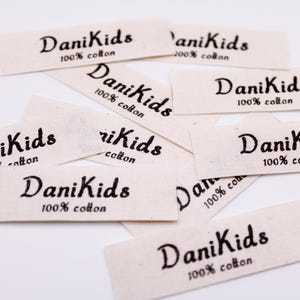 May include: Off-white fabric labels, each bearing the text "DaniKids" in a cursive script, and "100% cotton". The labels are scattered across a white surface, ideal for children's clothing.