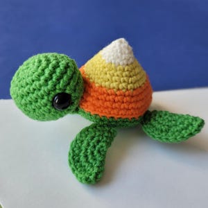 May include: A crocheted turtle toy with a candy corn shell. The turtle is green with black eyes and a white, yellow, and orange shell.