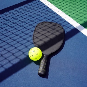 May include: A black pickleball paddle and a yellow pickleball on a blue and green pickleball court.