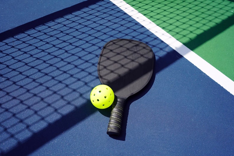 K&ouml;nnte beinhalten: Ein schwarzer Pickleball-Schl&auml;ger und ein gelber Pickleball auf einem blauen und gr&uuml;nen Pickleball-Platz.