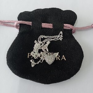 May include: A silver necklace with a heart-shaped pendant featuring a wing design and encrusted with small, sparkling stones. The necklace rests on a black velvet pouch with a pink drawstring. The pouch has the word "PANDORA" printed on it.