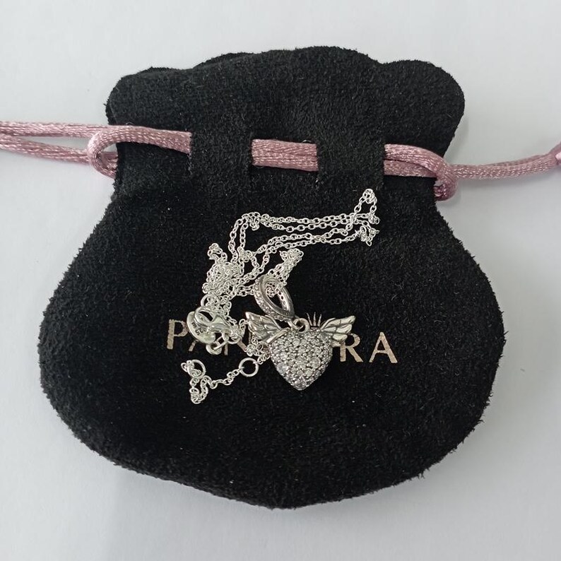May include: A silver necklace with a heart-shaped pendant featuring a wing design and encrusted with small, sparkling stones. The necklace rests on a black velvet pouch with a pink drawstring. The pouch has the word "PANDORA" printed on it.