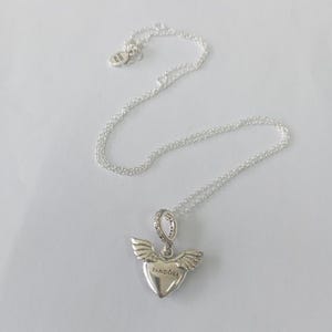 May include: A silver necklace with a heart-shaped pendant featuring wings and the word "Pandora". The pendant is embellished with small, sparkling stones. The necklace chain is delicate and lies on a white surface.
