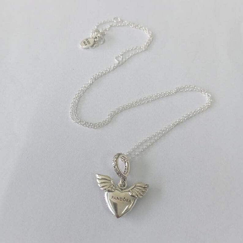 May include: A silver necklace with a heart-shaped pendant featuring wings and the word "Pandora". The pendant is embellished with small, sparkling stones. The necklace chain is delicate and lies on a white surface.